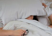 “밤에 자꾸 깨요”…수면장애의 뜻밖의 원인, ‘체온 조절 실패’ person lying on bed while covering face with pillow and holding eyeglasses