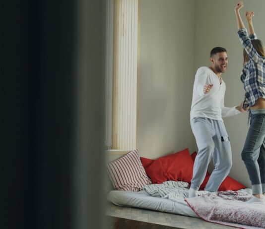 층간소음 걱정 없는 ‘방구석 댄스’ 홈트레이닝 꿀팁 Couple dancing joyfully on a bed near window.