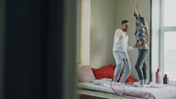 Couple dancing joyfully on a bed near window.